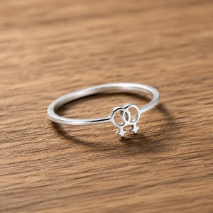 Op de afbeelding: Een zilveren ring met een dubbel vrouwelijk symbool. De ring is dun en delicaat, met het symbool als middelpunt. De ring ligt op een houten achtergrond.