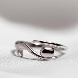 Op de afbeelding: Een zilverkleurige ring met een gestileerd kattenontwerp. De ring heeft een gebogen staart en een kattenkop met puntige oren. De ring is verstelbaar en ligt op een witte ondergrond.