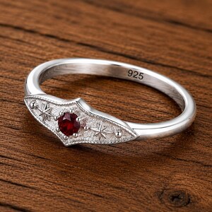 May include: A sterling silver ring featuring a central round-cut red gemstone set within a pointed, textured shield-shaped centerpiece. The design includes two engraved star motifs and small metal beads. The inner band is stamped with 925.