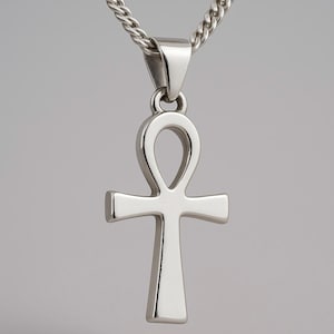 May include: A silver ankh pendant necklace. The ankh, a cross with a loop at the top, hangs from a silver chain. The pendant and chain are made of a shiny, reflective metal, likely sterling silver, against a neutral background.