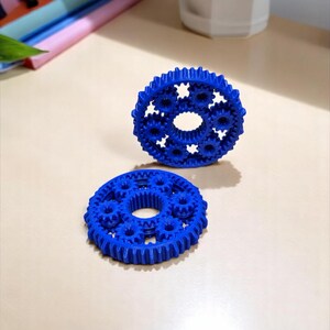 Può includere: Due modelli di ingranaggi blu sono esposti su una superficie beige chiaro. Ogni ingranaggio ha un foro centrale circondato da ingranaggi più piccoli. Gli ingranaggi sono realizzati in un materiale solido e opaco e sono posizionati per mostrare il loro design.