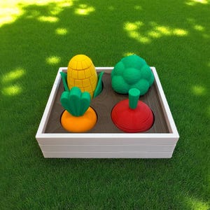 May include: A white wooden box filled with colorful, stylized vegetables. The box contains a yellow corn, green broccoli, orange carrot, and red beet. The vegetables are arranged in a square pattern, set against a green grass background.