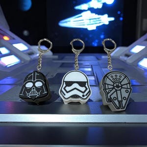 Può includere: Tre portachiavi con personaggi iconici di Star Wars: Darth Vader, uno Stormtrooper e il Millennium Falcon. Ogni portachiavi ha una catena e un anello argentati. Il portachiavi di Darth Vader è nero e bianco, lo Stormtrooper è bianco e nero e il Millennium Falcon è grigio.