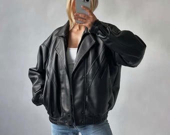 Puede incluir: Chaqueta de cuero negra con diseño tipo bomber. Presenta un cuello ancho, cierre de cremallera y múltiples bolsillos. Las mangas son largas y fruncidas en los puños. La chaqueta se lleva con un top blanco y vaqueros azules.