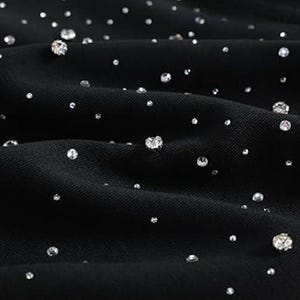 May include: Close-up of black fabric adorned with scattered, sparkling rhinestones of varying sizes. The rhinestones catch the light, creating a shimmering effect against the dark background. The fabric is slightly textured, with soft folds.