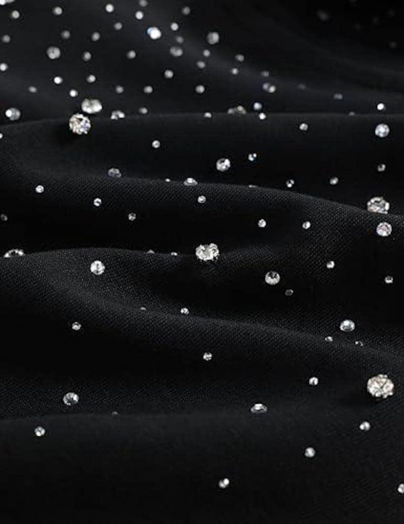May include: Close-up of black fabric adorned with scattered, sparkling rhinestones of varying sizes. The rhinestones catch the light, creating a shimmering effect against the dark background. The fabric is slightly textured, with soft folds.