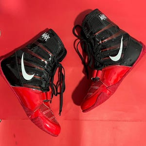 May include: Pair of high-top wrestling shoes. The shoes are primarily black and red, with a glossy finish on the red sections. White Nike swooshes are visible on the sides. The shoes have laces and a strap closure. The background is a solid red.