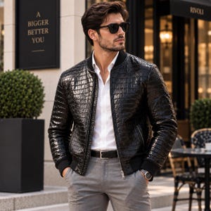 Pode incluir: Um blusão bomber preto com textura brilhante em relevo de crocodilo, apresentando gola, punhos e bainha canelados. O casaco é usado sobre uma camisa branca de botões, combinada com calças cinzentas e um cinto preto para um visual estruturado.