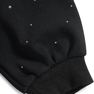 May include: Close-up of a black sweatshirt sleeve with scattered, small, shiny rhinestones. The cuff is ribbed and dark, contrasting with the smooth fabric above. The garment appears to be a casual top.