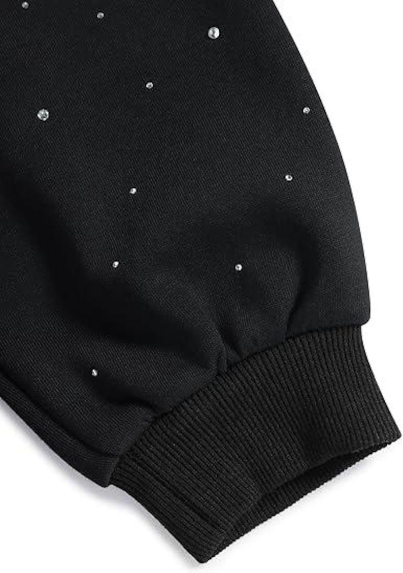 May include: Close-up of a black sweatshirt sleeve with scattered, small, shiny rhinestones. The cuff is ribbed and dark, contrasting with the smooth fabric above. The garment appears to be a casual top.