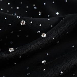 May include: Close-up of black fabric adorned with numerous sparkling, silver-colored rhinestones of varying sizes. The fabric is draped, creating soft folds and shadows. The rhinestones are scattered across the surface, catching the light and creating a shimmering effect.