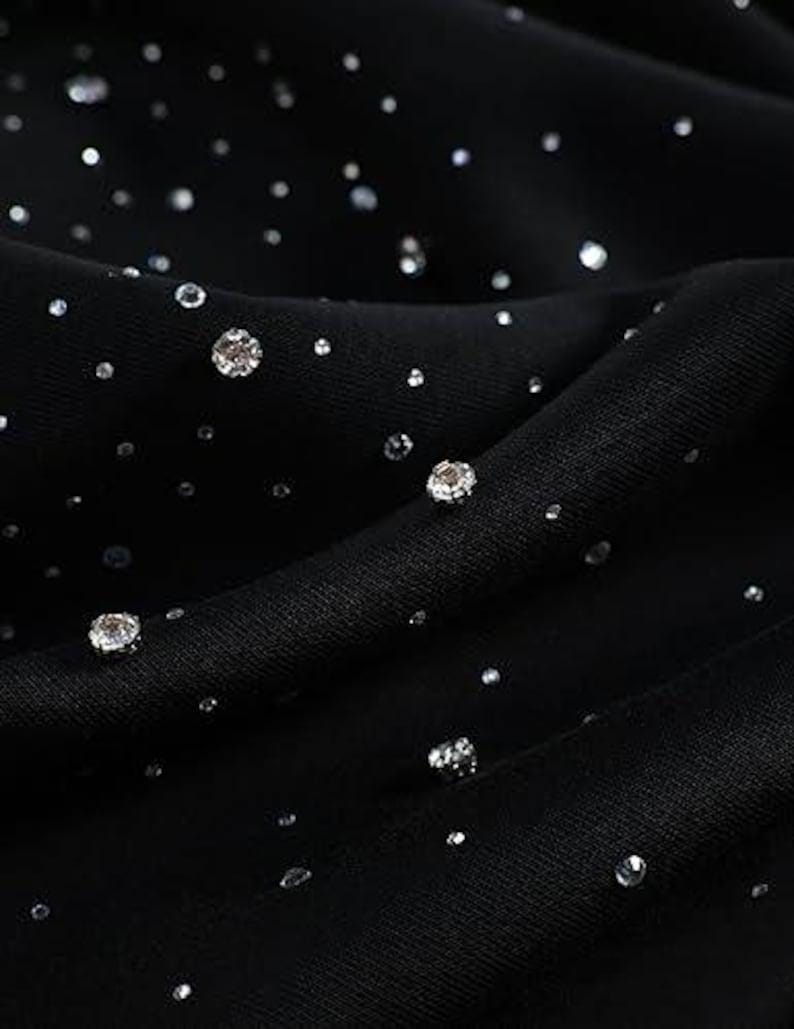 May include: Close-up of black fabric adorned with numerous sparkling, silver-colored rhinestones of varying sizes. The fabric is draped, creating soft folds and shadows. The rhinestones are scattered across the surface, catching the light and creating a shimmering effect.