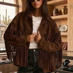 May include: A brown suede fringe jacket with long fringe detailing, worn over a white top and black leather trousers. The background shows pottery and jewellery displays. The jacket is a key fashion item.