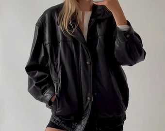 May include: Black leather jacket with a button-up front, side pockets, and a collared neckline. The jacket has a slightly oversized fit and a ribbed hem. The sleeves are long and have buttoned cuffs. The jacket is worn over a white shirt.