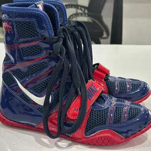 May include: Pair of blue and red Nike boxing shoes with white swoosh logos. The high-top shoes have mesh panels and red straps with the Nike logo. The laces are black.