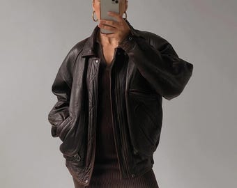 May include: A dark brown leather jacket with a classic bomber style. The jacket features a collared neckline, front pockets, and a ribbed hem. The jacket is worn over a brown ribbed dress.
