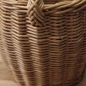 May include: A light brown woven basket featuring a tight, intricate wicker pattern. The structure displays a consistent, repeating weave design with a visible handle attachment point at the top rim, showcasing the natural texture of the material.
