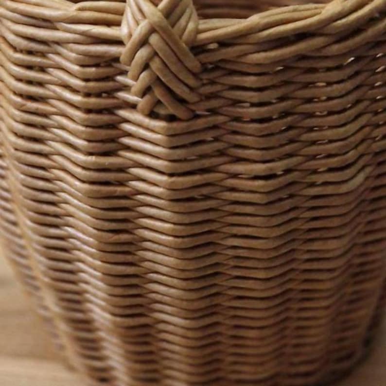 May include: A light brown woven basket featuring a tight, intricate wicker pattern. The structure displays a consistent, repeating weave design with a visible handle attachment point at the top rim, showcasing the natural texture of the material.