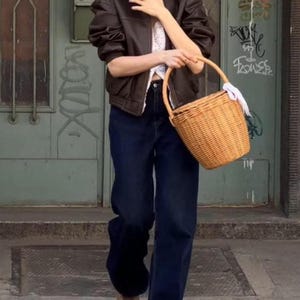 May include: A person wearing a brown leather jacket, white top, and dark blue jeans, holding a woven basket with a white cloth. The background features a green door with graffiti.