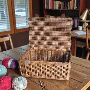 May include: A woven wicker basket with an open lid, showcasing its interior. The basket is a warm brown color, with leather straps and clasps. Several balls of yarn in shades of red, purple, and teal are on the wooden table next to the basket. Knitting needles are also visible.