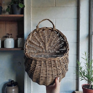 Pode incluir: Um cesto de parede de vime castanho tecido com uma pega. O cesto tem uma forma arredondada e uma parte traseira plana, projetada para pendurar numa parede. O cesto está vazio e parece novo. O cesto é segurado em frente a uma parede branca.