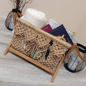 May include: A wooden magazine rack with a macrame front, holding books and a white knitted item. The rack has a natural wood finish and a woven design. Decorative items, including a black wire basket, are in the background.