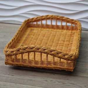 May include: A rectangular, woven wicker basket with a curved handle. The basket is light brown and has a braided border. The interior is a flat surface. The basket is likely used for serving or storage.