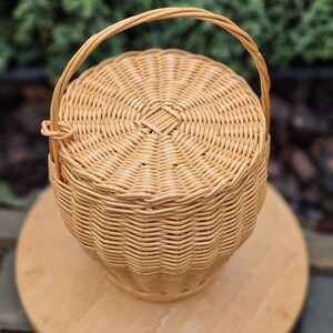May include: A round, light tan woven wicker basket with a matching lid and a single arched handle. The basket features a tight, circular weave pattern on the lid and a vertical, textured weave along the sides, creating a structured, cylindrical shape.