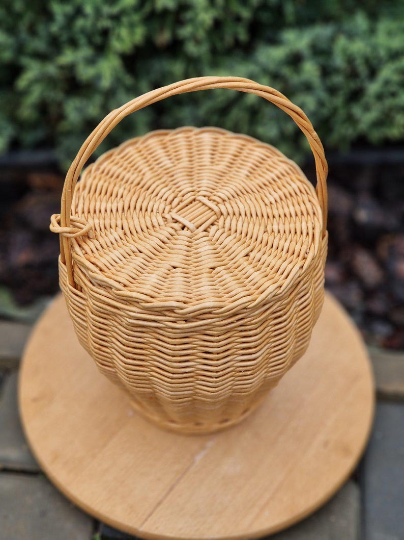 May include: A round, light tan woven wicker basket with a matching lid and a single arched handle. The basket features a tight, circular weave pattern on the lid and a vertical, textured weave along the sides, creating a structured, cylindrical shape.