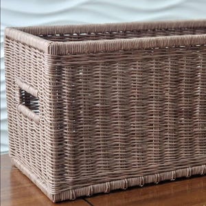 May include: A rectangular brown woven basket with a handle on each side. The basket is made of tightly woven material and has a decorative trim along the bottom edge. The basket is sitting on a wooden surface.