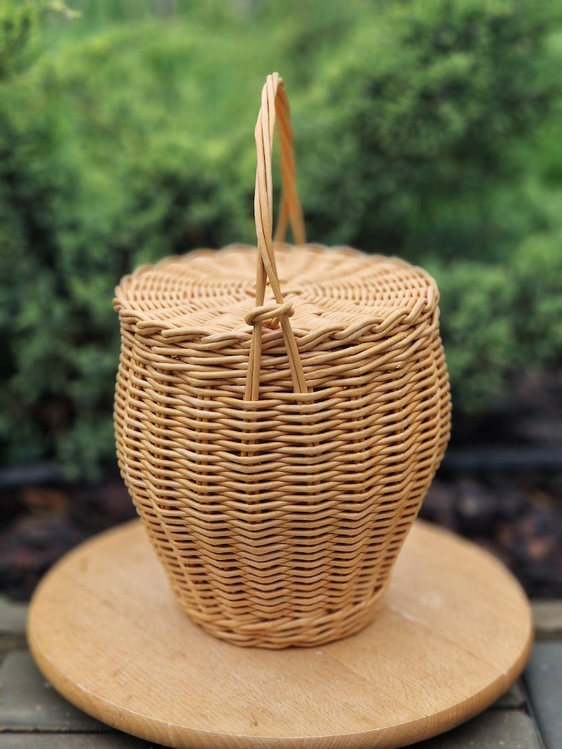 May include: A light brown woven wicker basket with a round lid and a single upright handle. The basket features a tapered cylindrical body with a tight, horizontal weave pattern and a braided edge detail around the top rim and the lid circumference.