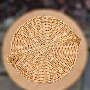 May include: A circular, light tan woven wicker basket viewed from directly above. The basket features a tight, concentric circular weave pattern and a single, thin, arched handle that extends across the diameter of the top opening.