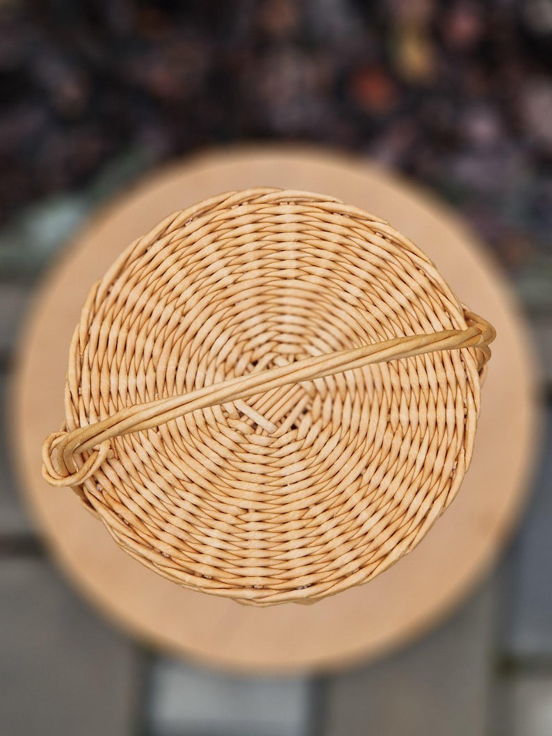 May include: A circular, light tan woven wicker basket viewed from directly above. The basket features a tight, concentric circular weave pattern and a single, thin, arched handle that extends across the diameter of the top opening.