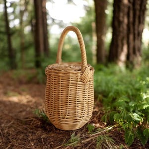 May include: A light brown woven wicker basket with a cylindrical body and a single arched handle. The basket features a tight, vertical weave pattern and a rounded lid that sits flush with the top rim, creating a structured and functional container.