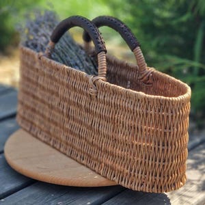 May include: An elongated oval woven basket with a light brown natural finish. The basket features two sturdy handles wrapped in dark brown leather with visible stitching. Dried lavender sprigs are tucked inside the open top of the basket.