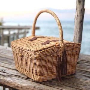 Peut inclure: Un panier de pique-nique rectangulaire en osier tressé avec une anse arquée unique. Le panier dispose d'un couvercle à charnière fixé par une petite fermeture à bouton en bois et boucle, orné de lanières en cuir marron et deux pompons en cuir.