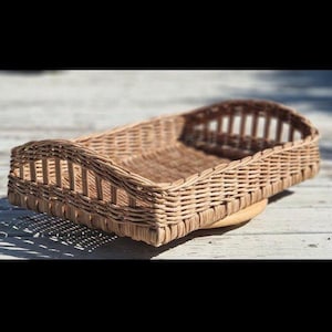 May include: A rectangular woven wicker basket with a light brown natural finish. The basket features a flat base and low, open-weave sides with vertical slats along the perimeter, creating a structured, tray-like appearance for holding items.