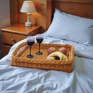 May include: A woven wicker serving tray with two wine glasses filled with red wine, apples, and a banana on a bed with light blue sheets. A wooden nightstand with a lamp is in the background.