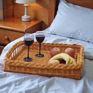 May include: A rectangular light brown woven wicker tray holding two stemmed wine glasses filled with dark red liquid, two round red apples, and a bunch of yellow bananas, all resting on a white bed sheet with a wooden headboard in the background.