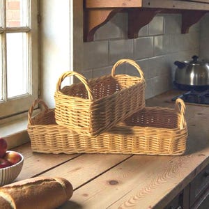 May include: Two woven wicker baskets with handles, one rectangular and one square, stacked on a wooden countertop. The baskets are a light tan color and appear to be empty. A loaf of bread and a bowl of apples are in the foreground.
