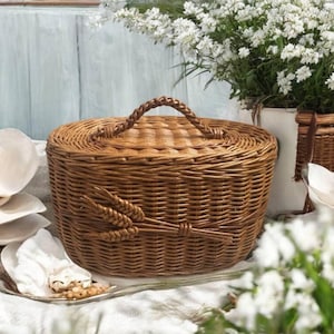 Può includere: Un cesto rotondo in vimini marrone con manico intrecciato e coperchio abbinato. Il cesto è decorato con un motivo a spiga di grano sul davanti. Il cesto è appoggiato su un panno bianco.