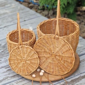 May include: Two round, light brown woven wicker baskets with matching lids and tall, upright handles. The baskets feature a circular base and a tight, concentric weave pattern. Several small, round, light-colored wooden beads are scattered.