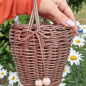 May include: A brown woven basket with a tapered shape and a single handle. The basket features a decorative brown cord tied in a bow at the front, with two light-colored wooden beads hanging from the ends of the cord strings.