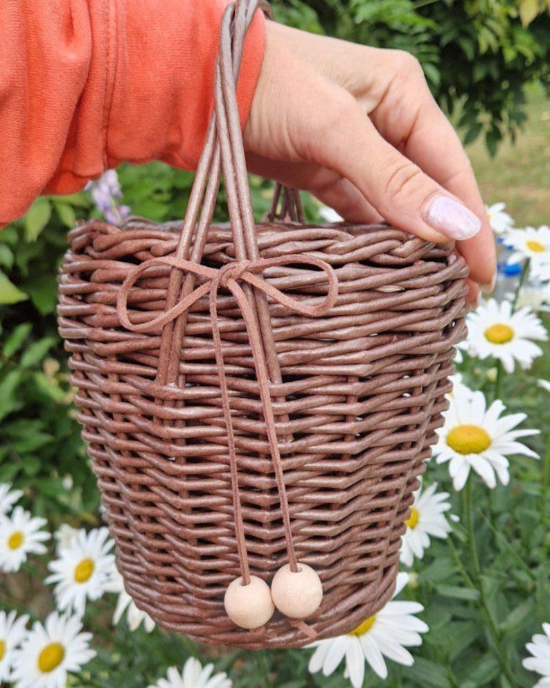 May include: A brown woven basket with a tapered shape and a single handle. The basket features a decorative brown cord tied in a bow at the front, with two light-colored wooden beads hanging from the ends of the cord strings.