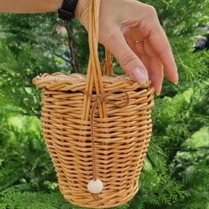 May include: A light brown woven wicker basket with a round lid and a tall vertical handle. A thin leather cord is tied around the handle, featuring a single round wooden bead at the end. The basket has a tapered, cylindrical shape with a textured finish.