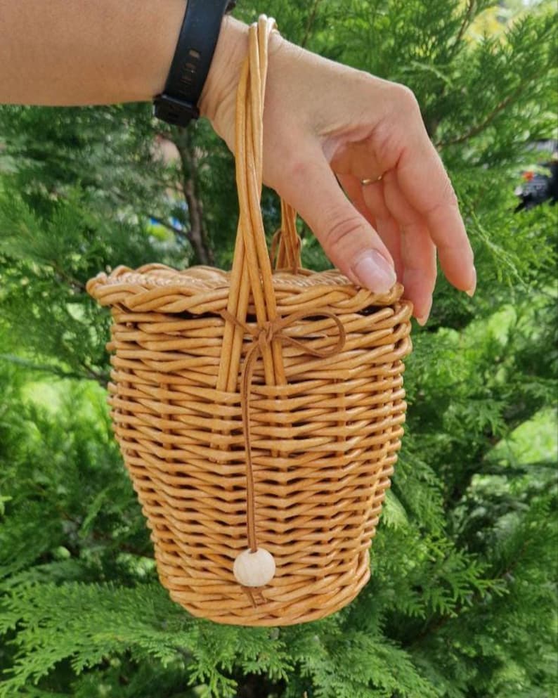 May include: A light brown woven wicker basket with a round lid and a tall vertical handle. A thin leather cord is tied around the handle, featuring a single round wooden bead at the end. The basket has a tapered, cylindrical shape with a textured finish.