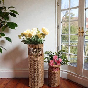 Może przedstawiać: Dwa tkane wazony na kwiaty z wikliny. Wyższy wazon zawiera żółte kwiaty, a krótszy różowe. Wazony są cylindryczne z dekoracyjnym wzorem tkanym w pobliżu góry. Wazony stoją na drewnianej podłodze.