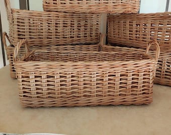 May include: A collection of rectangular woven wicker baskets featuring a natural light brown finish. Each basket has a sturdy base, vertical side walls with a consistent cross-weave pattern, and two arched handles attached to the short ends.