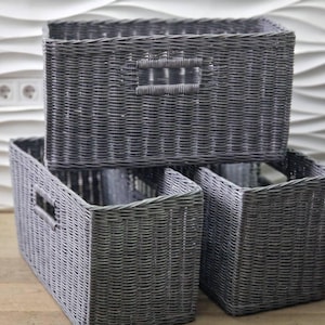 May include: Three gray woven storage baskets stacked together. The baskets are rectangular with cut-out handles. The baskets are made of a woven material and are a solid gray color. The background is a white wall with a wavy pattern.