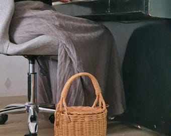 May include: A light brown, cylindrical woven wicker basket with a flat lid and a single arched handle. The basket features a tight, vertical weave pattern and a small wooden bead detail attached to the side near the top rim of the structure.